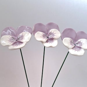 Pode incluir: Tr&ecirc;s flores de amor-perfeito em cer&acirc;mica, com p&eacute;talas lavanda e centros brancos. Montadas em hastes de arame verde, apresentam detalhes de veios e um acabamento brilhante. Uma decora&ccedil;&atilde;o floral delicada e art&iacute;stica.