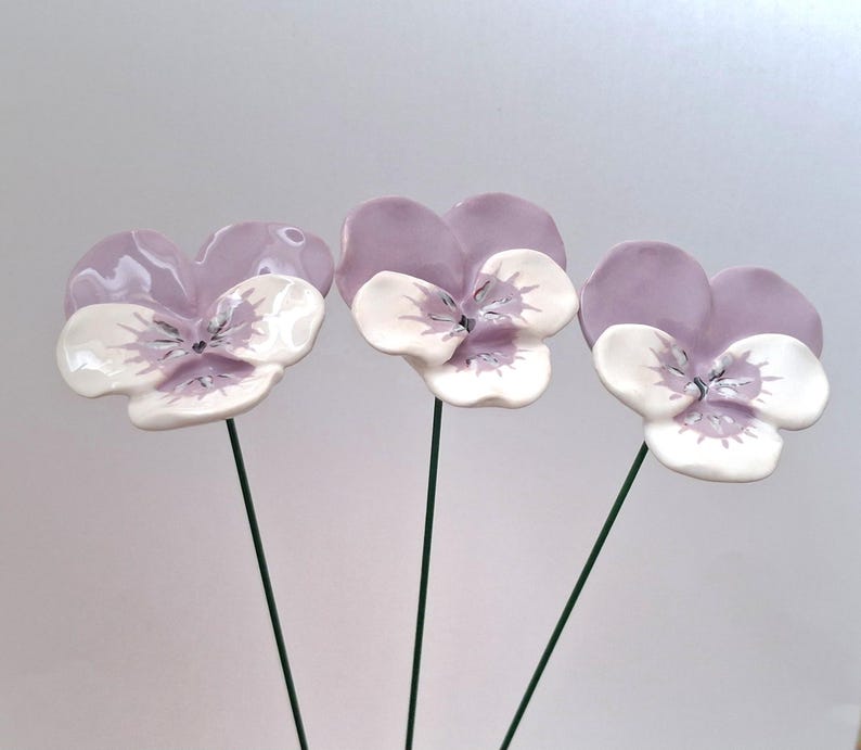 Pode incluir: Tr&ecirc;s flores de amor-perfeito em cer&acirc;mica, com p&eacute;talas lavanda e centros brancos. Montadas em hastes de arame verde, apresentam detalhes de veios e um acabamento brilhante. Uma decora&ccedil;&atilde;o floral delicada e art&iacute;stica.