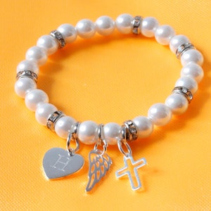 Op de afbeelding: Een witte parel armband met zilveren bedels, waaronder een hart met de letter H, een vleugel en een kruis.