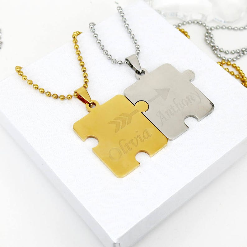May include: Two gold and silver puzzle piece necklaces with the names Olivia and Anthony engraved on them. The puzzle pieces are connected by an arrow symbol.