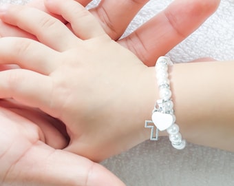 May include: A white pearl bracelet with a silver heart charm and a cross pendant. The bracelet is on a child's wrist.