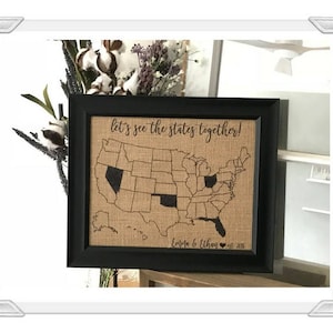 May include: A black framed burlap map of the United States with the text "Let's see the states together!" and "Emily & Ethan" with a heart and the year 2018.