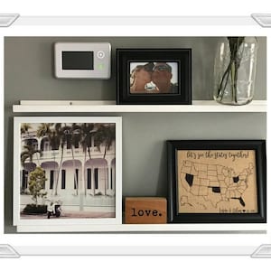 May include: A white shelf with four framed pictures. The top left picture is a black and white photo of a couple. The top right picture is a black and white photo of a couple. The bottom left picture is a color photo of a building with palm trees. The bottom right picture is a burlap map of the United States with the text "Let's see the states together!" and the text "Love." is below the map.