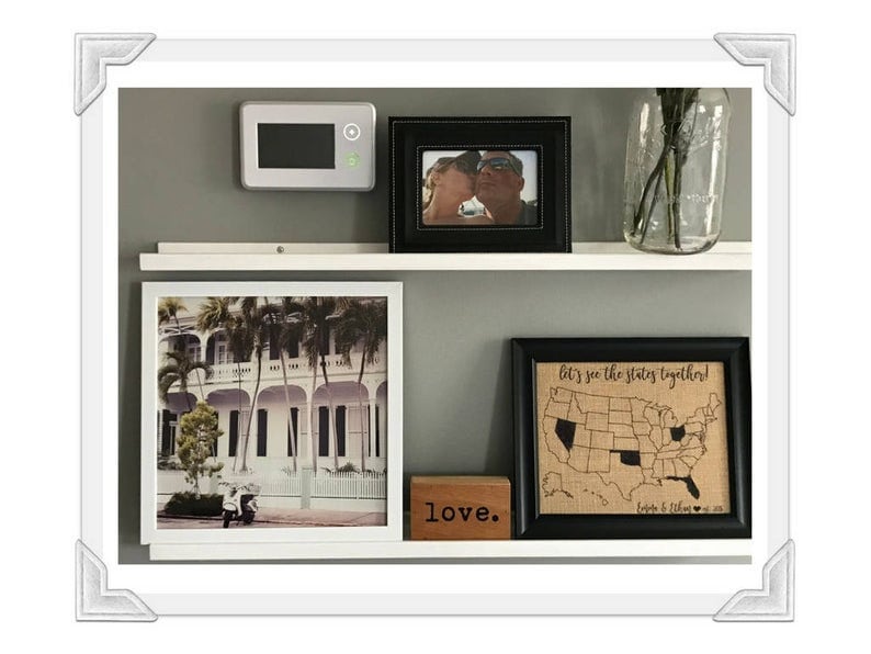 May include: A white shelf with four framed pictures. The top left picture is a black and white photo of a couple. The top right picture is a black and white photo of a couple. The bottom left picture is a color photo of a building with palm trees. The bottom right picture is a burlap map of the United States with the text "Let's see the states together!" and the text "Love." is below the map.
