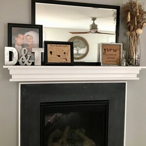 May include: A white mantelpiece with a black fireplace below. The mantelpiece is decorated with a framed photo, a framed map of the United States, a wooden sign that says "grow old with me, the best is yet to be", and a metal sign that says "D&T".