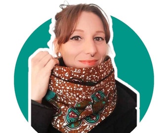 May include: A brown and teal patterned cowl neck warmer. The fabric features a repeating floral design with white accents. The cowl is worn over a black garment, creating a layered look. The background is a teal circle.