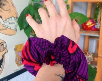 May include: A vibrant purple and red patterned scrunchie. The scrunchie is wrapped around a wrist, showcasing its bold colors and design. The background includes a vintage anatomical chart and greenery.