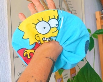 May include: A blue sleep mask featuring a yellow cartoon character with large eyes and spiky yellow hair. The character's face is centered on the mask, with a red and yellow design at the bottom. The mask is held in a hand.