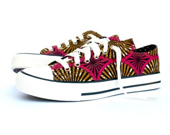 May include: Pair of canvas sneakers with a vibrant pink and gold geometric pattern. The shoes have white laces, a white rubber sole, and a black stripe. The design is eye-catching and stylish.