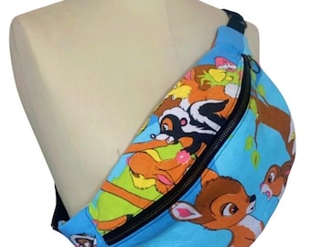 May include: A blue fanny pack featuring a colorful print of Bambi and other forest animals. The bag has a black zipper and strap, and is displayed on a mannequin. The design includes images of a skunk, deer, and other characters.