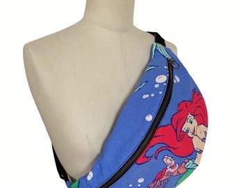 May include: Blue fanny pack featuring a colorful graphic of Ariel from The Little Mermaid. The bag has a black zipper and strap. The design includes bubbles and other characters from the movie. The fanny pack is displayed on a mannequin.