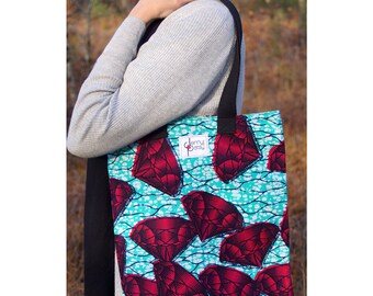 May include: A tote bag with a vibrant pattern of large red diamonds on a turquoise background. The bag has black straps and a label that reads "Jenny Dotsy". The bag is held by a person.