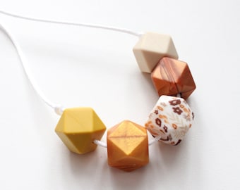 May include: A necklace made of six geometric silicone beads in various colors and patterns. The beads are strung on a white cord. The beads are yellow, beige, brown, and white with a floral pattern.