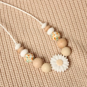 May include: A white and beige silicone and wooden bead necklace with a white daisy charm. The necklace is on a beige knitted surface.