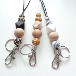 May include: Three keychains with wooden beads and different patterned beads. The beads are in black, white, brown, and orange. Each keychain has a silver metal ring and a silver metal clip.