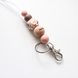 May include: A white and brown patterned wooden bead with the name "Jenn" engraved on it, a brown bead, a pink bead, and a silver key chain with a clip.