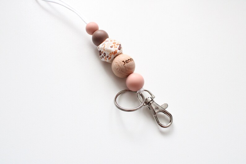 May include: A white and brown patterned wooden bead with the name "Jenn" engraved on it, a brown bead, a pink bead, and a silver key chain with a clip.