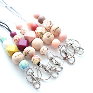 May include: Five colorful beaded lanyards with personalized wooden beads and silver swivel clips. The beads are in shades of pink, brown, yellow, white, and blue. The wooden beads have the names "Jenny", "Miss Smith", "Miss Brogan", and "Jolande" engraved on them.