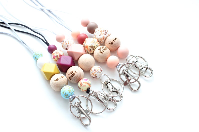 May include: Five colorful beaded lanyards with personalized wooden beads and silver swivel clips. The beads are in shades of pink, brown, yellow, white, and blue. The wooden beads have the names "Jenny", "Miss Smith", "Miss Brogan", and "Jolande" engraved on them.