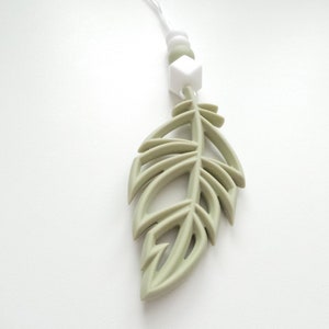 May include: A sage green silicone feather pendant with a white silicone bead and a white silicone cube charm. The pendant is attached to a white cord.