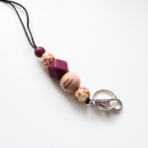 May include: A burgundy and cream colored beaded lanyard with a wooden bead that says "Miss Brogan" and a silver key ring.
