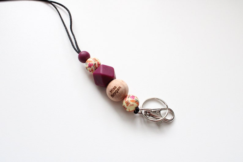 May include: A burgundy and cream colored beaded lanyard with a wooden bead that says "Miss Brogan" and a silver key ring.