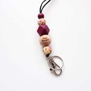 May include: A burgundy and cream floral patterned lanyard with a wooden bead that says "Miss Brogan" and a silver key chain.