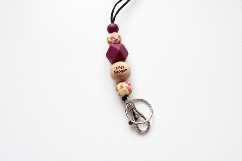 May include: A burgundy and cream floral patterned lanyard with a wooden bead that says "Miss Brogan" and a silver key chain.