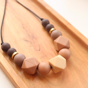 May include: A necklace featuring a black cord and a selection of brown beads. The beads are round and geometric, in various shades of brown, with gold spacers. The necklace is presented on a wooden tray.