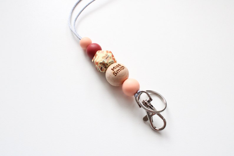 May include: A light pink, red, and brown beaded lanyard with a wooden bead that says "Miss Smith" and a silver swivel clasp.