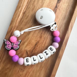 May include: A baby's dummy clip with the name "ELLIE" spelled out in white letter beads. The clip features a butterfly charm and a string of pink, purple, and white beads. The dummy clip is attached to a white dummy.