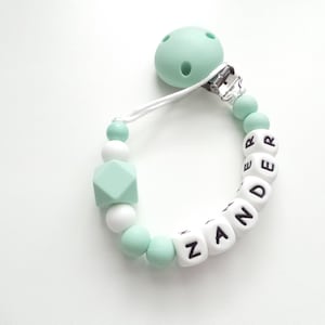 May include: A mint green and white silicone baby teether with a clip and the name "ZANDER" spelled out in black letters.
