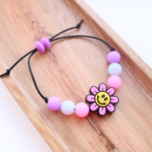 May include: A pink and purple beaded bracelet with a pink flower charm featuring a yellow smiley face. The bracelet is made with silicone beads and has a black adjustable cord.