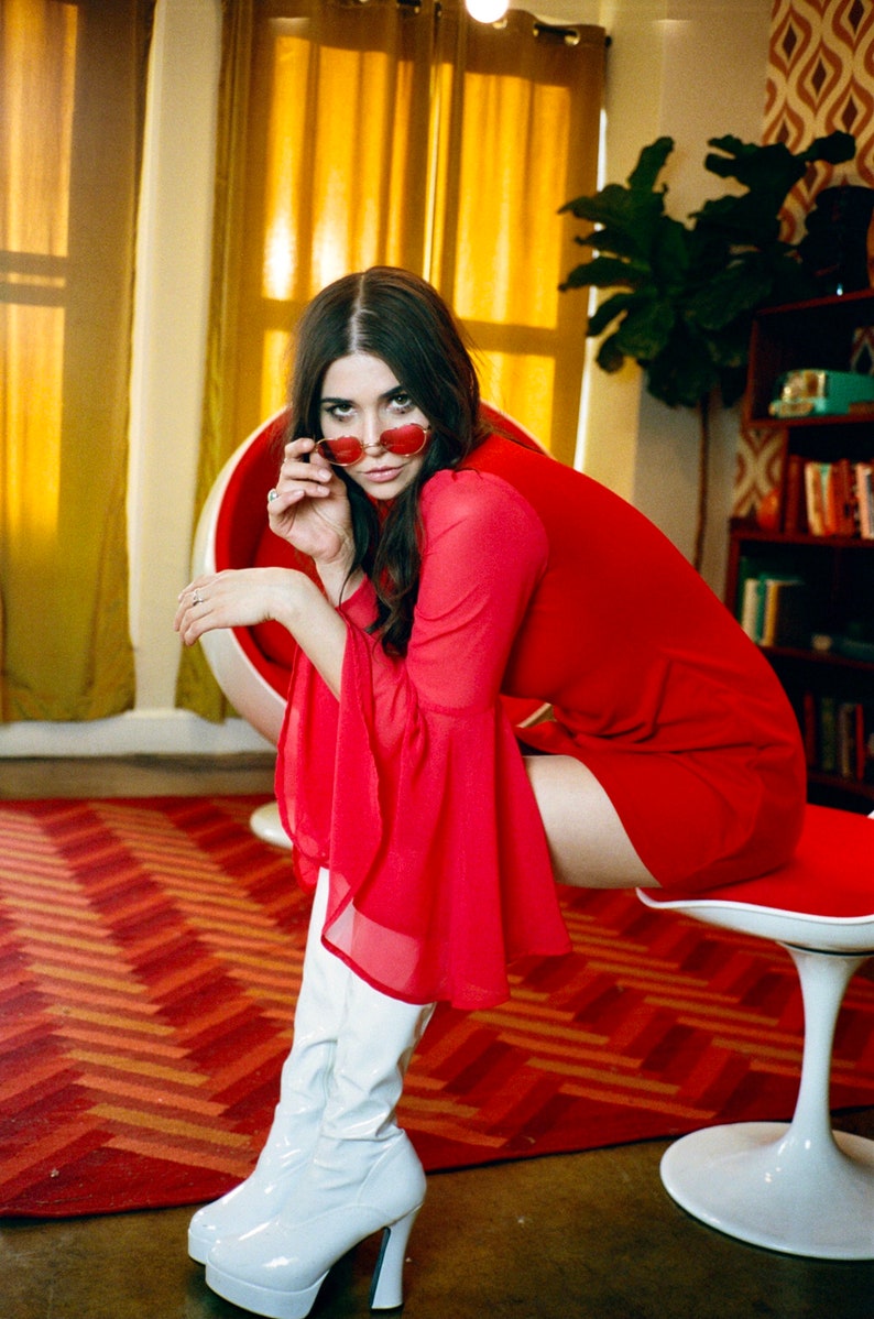 Puede incluir: Una persona con un vestido rojo con mangas acampanadas y botas blancas de plataforma. Sostiene gafas de sol rojas en forma de coraz&oacute;n. El escenario incluye una silla roja y blanca, una alfombra estampada y una estanter&iacute;a.