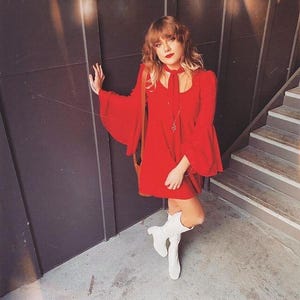 Puede incluir: Un vestido rojo brillante con mangas acampanadas y un lazo a juego en el cuello. El vestido se combina con botas blancas y un bolso de hombro marr&oacute;n. El conjunto se encuentra sobre un fondo oscuro con una escalera.