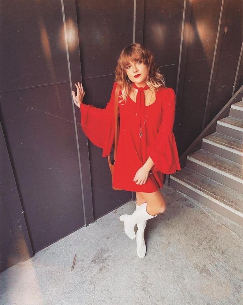Puede incluir: Un vestido rojo brillante con mangas acampanadas y un lazo a juego en el cuello. El vestido se combina con botas blancas y un bolso de hombro marr&oacute;n. El conjunto se encuentra sobre un fondo oscuro con una escalera.