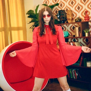 Puede incluir: Vestido rojo con mangas acampanadas y lazo en el cuello. El atuendo incluye botas blancas hasta la rodilla y gafas de sol en forma de coraz&oacute;n. El fondo muestra un interior retro con una silla huevo y papel pintado estampado.