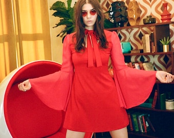 Pode incluir: Um vestido curto vermelho vibrante com mangas compridas e largas, e um decote alto com detalhe de laço. O vestido tem um corpete justo que se alarga ligeiramente na bainha, criando uma silhueta estruturada. Combinado com botas brancas de cano alto.
