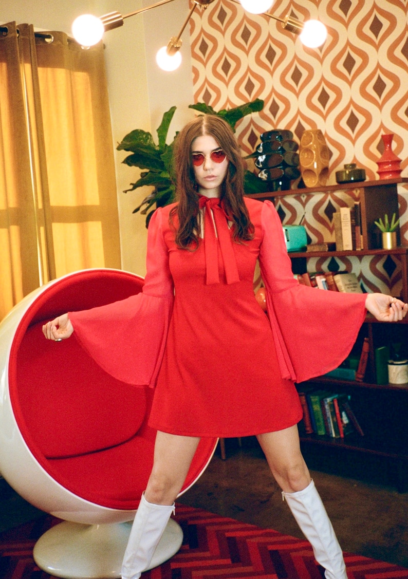 Puede incluir: Vestido rojo con mangas acampanadas y lazo en el cuello. El atuendo incluye botas blancas hasta la rodilla y gafas de sol en forma de coraz&oacute;n. El fondo muestra un interior retro con una silla huevo y papel pintado estampado.
