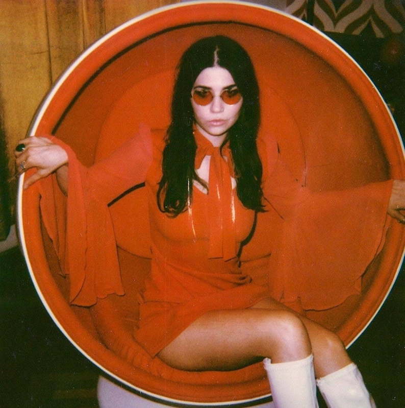 Puede incluir: Una persona con un vestido rojo y gafas de sol est&aacute; sentada en una gran silla redonda y naranja. El vestido tiene mangas largas y fluidas y un lazo en el cuello. Lleva botas blancas. El fondo es c&aacute;lido y de estilo retro.