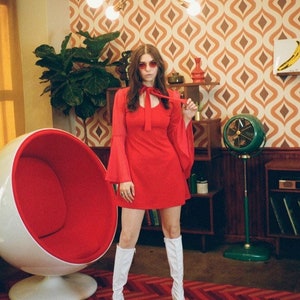 Puede incluir: Una persona con un vestido rojo y botas blancas est&aacute; en una habitaci&oacute;n retro. El vestido tiene mangas largas y acampanadas y un lazo en el cuello. La habitaci&oacute;n cuenta con una silla huevo roja y blanca, un ventilador verde y papel pintado estampado.