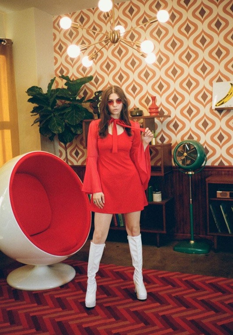 Puede incluir: Una persona con un vestido rojo y botas blancas est&aacute; en una habitaci&oacute;n retro. El vestido tiene mangas largas y acampanadas y un lazo en el cuello. La habitaci&oacute;n cuenta con una silla huevo roja y blanca, un ventilador verde y papel pintado estampado.