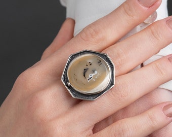 Può includere: Un anello d'argento con una montatura ottagonale che presenta una pietra di agata beige con inclusiones nere. L'anello è indossato su una mano con smalto per unghie marrone chiaro.