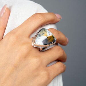 Puede incluir: Un anillo de plata con una piedra triangular grande. La piedra presenta un patr&oacute;n &uacute;nico de blanco, amarillo y gris. El anillo se lleva en un dedo, con tela blanca en el fondo. El anillo es una joya.