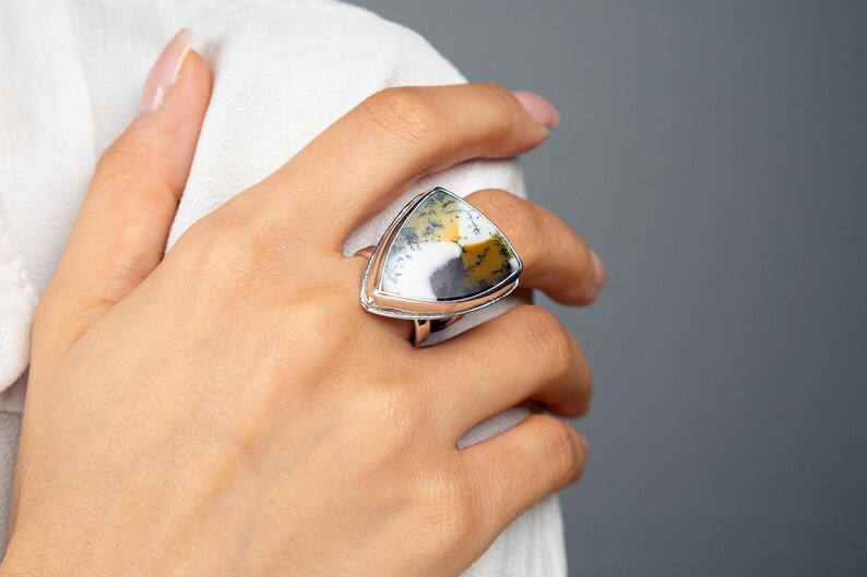 Puede incluir: Un anillo de plata con una piedra triangular grande. La piedra presenta un patr&oacute;n &uacute;nico de blanco, amarillo y gris. El anillo se lleva en un dedo, con tela blanca en el fondo. El anillo es una joya.