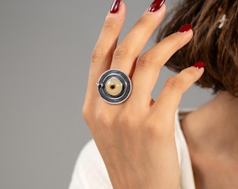 Puede incluir: Un anillo de plata con un diseño circular grande. El anillo presenta una piedra central de color claro rodeada por una banda gris oscuro y un anillo exterior texturizado. El anillo se lleva en una mano con esmalte de uñas rojo.