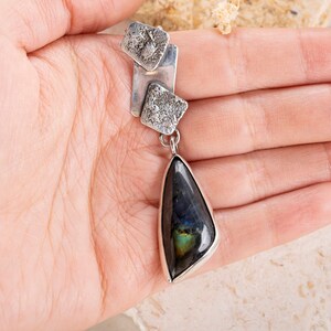 May include: A silver pendant necklace with a teardrop-shaped labradorite gemstone. The pendant features three silver squares with textured surfaces.