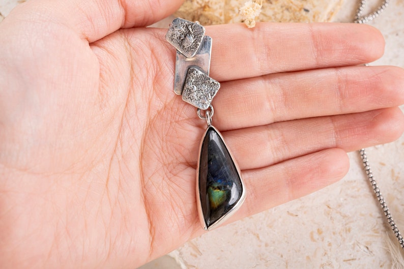 May include: A silver pendant necklace with a teardrop-shaped labradorite gemstone. The pendant features three silver squares with textured surfaces.