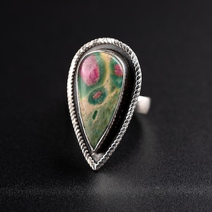 Puede incluir: Un anillo de plata con una piedra en forma de lágrima. La piedra presenta tonos verdes y beige con manchas rojas. El anillo tiene un borde negro y un diseño plateado en forma de cuerda alrededor de la piedra.