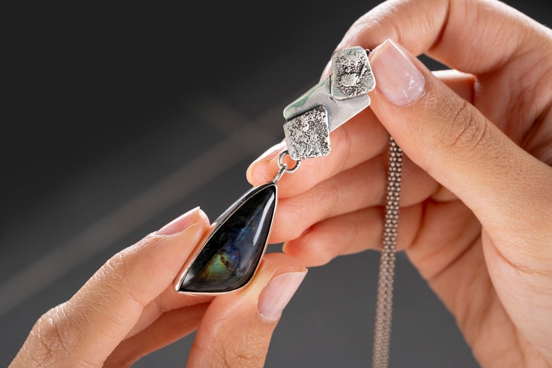 May include: A silver necklace with a textured pendant featuring a dark iridescent gemstone. The pendant is shaped like a teardrop and hangs from a delicate silver chain.